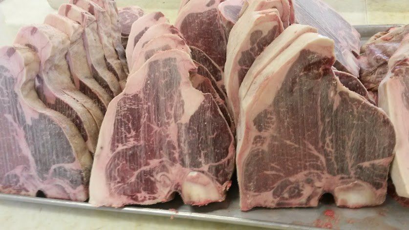 A tray holding several rows of raw T-bone steaks with visible fat marbling and bone segments.