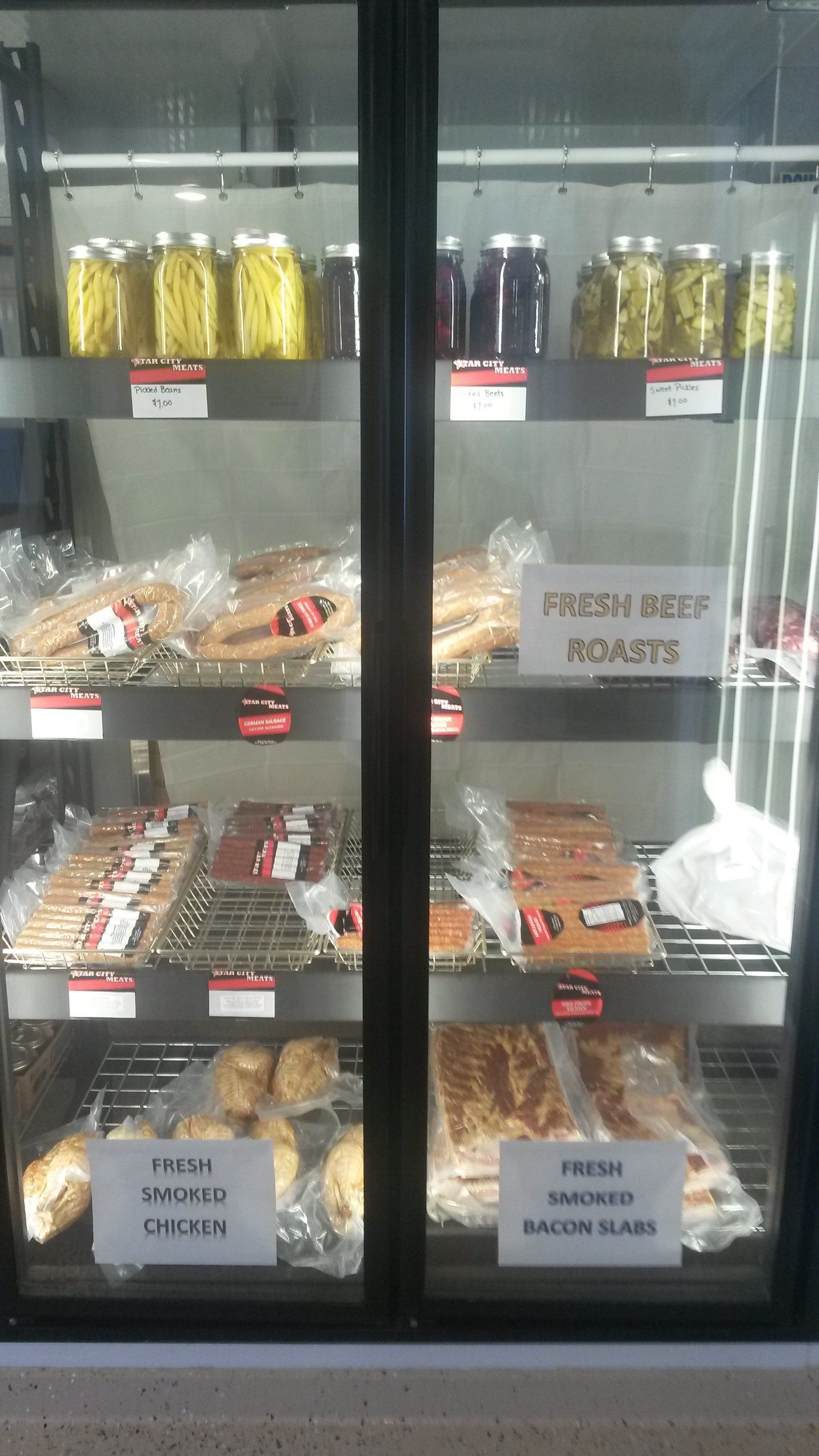 A grocery display case with shelves of jarred goods, bread, sausages, smoked cheeses, and bacon slabs.