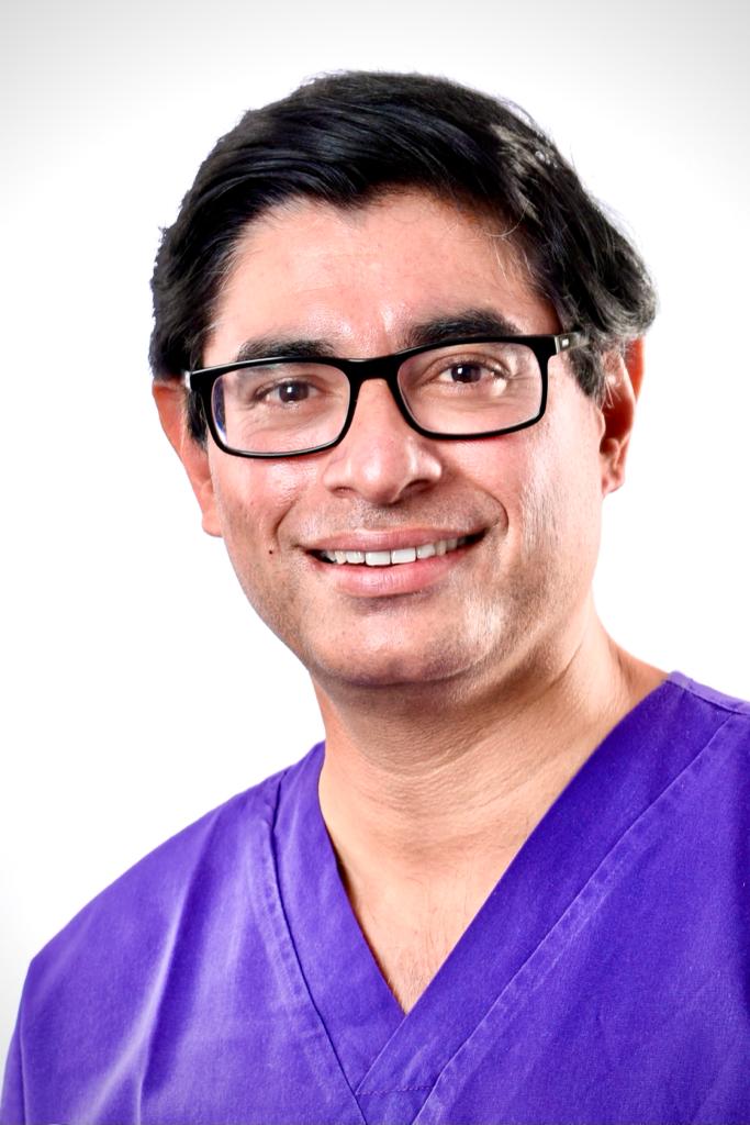 A man wearing glasses and a purple scrub top smiles for the camera.