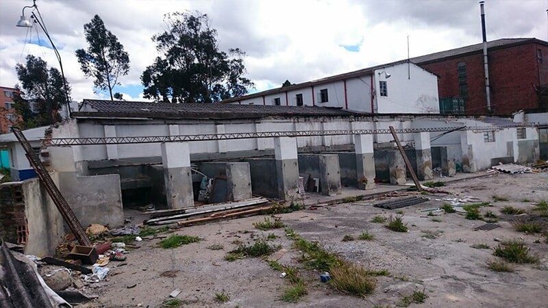 Patio de hormigón abandonado con maleza crecida y una hilera de antiguos edificios industriales bajo un cielo nublado.