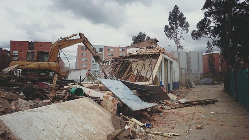 Excavadora de demolición junto a un edificio derrumbado y escombros esparcidos en un terreno polvoriento.