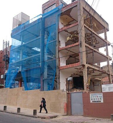 Edificio en obras rodeado de una red de seguridad azul, con un peatón caminando junto a una acera vallada.