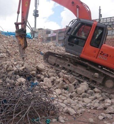 Excavadora naranja sobre un montón de escombros en una obra de demolición con barras de refuerzo retorcidas en primer plano.