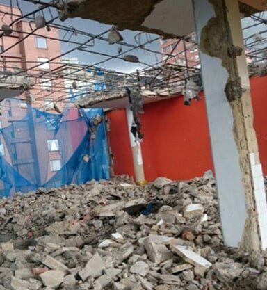 Habitación parcialmente demolida con escombros, columnas de hormigón a la vista, red azul y paredes rojas.