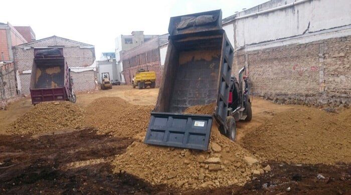 Camiones volquete descargando tierra en una obra en construcción fangosa junto a muros de ladrillo.