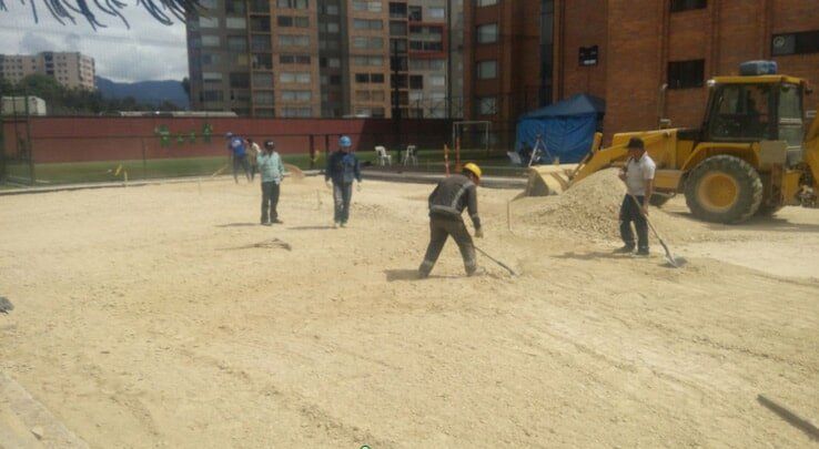 Trabajadores nivelando un terreno arenoso junto a un edificio de apartamentos y un tractor amarillo.