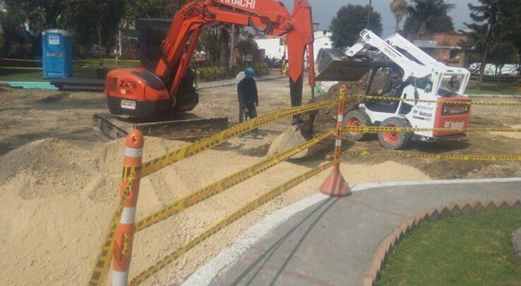 Obra en construcción con excavadora naranja y minicargadora blanca junto a una excavación arenosa vallada.