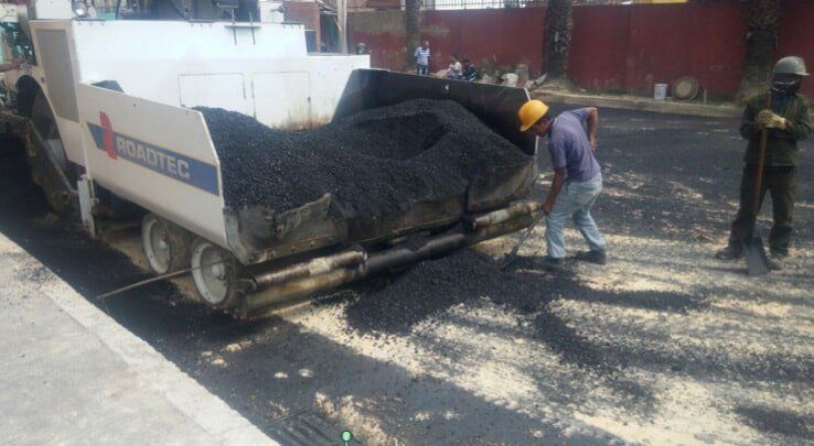 Trabajadores pavimentando una carretera con asfalto negro desde un camión; uno de ellos palea cerca del material esparcido.