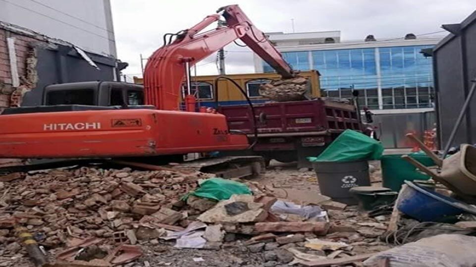 Una excavadora naranja retira escombros en una obra en construcción junto a un camión rojo dañado.
