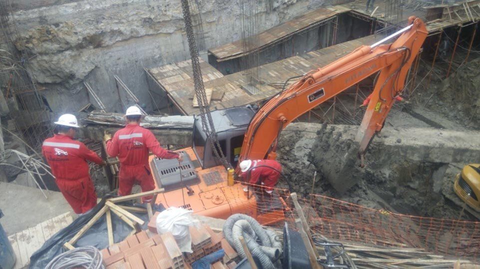 Una excavadora naranja en una zanja profunda, con dos trabajadores con trajes de seguridad rojos cerca.