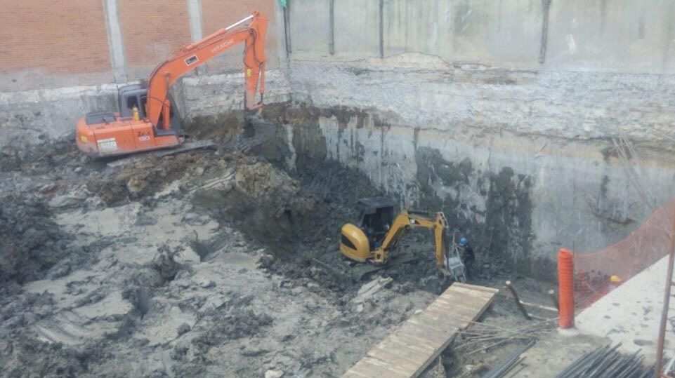 Excavadora en una profunda fosa de construcción con trabajadores y terreno fangoso, paredes de hormigón a su alrededor.
