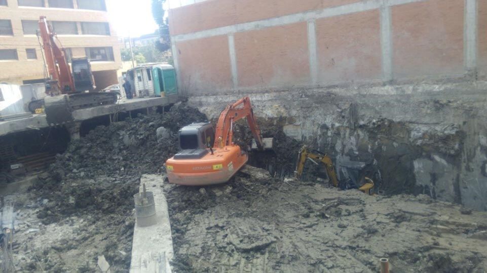 Excavadoras removiendo tierra fangosa en una obra de construcción junto a un muro de ladrillos.