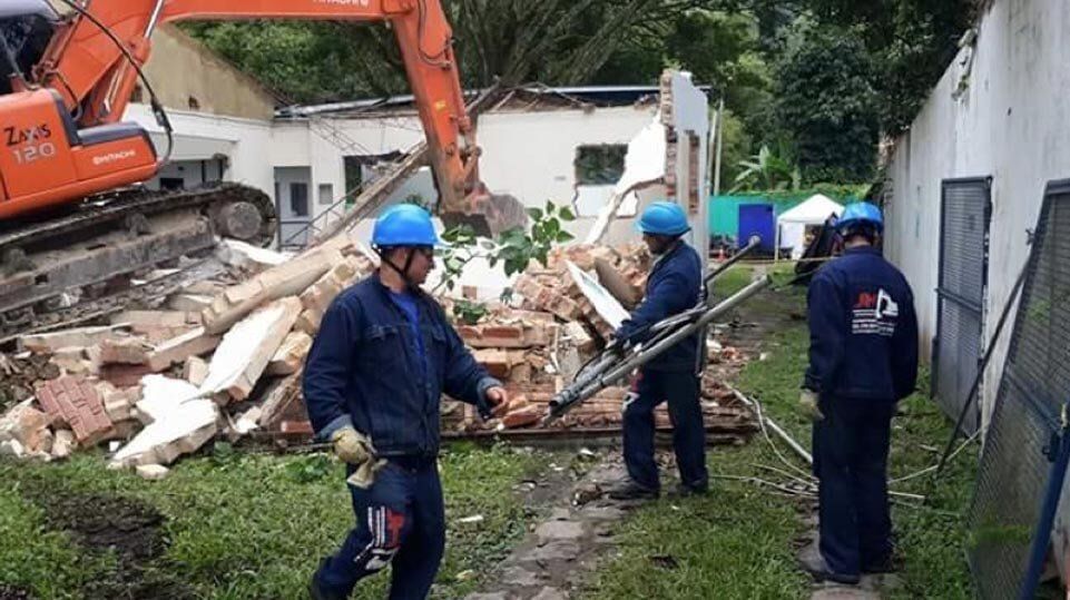 Trabajadores con cascos azules retiran escombros junto a un muro derrumbado y una excavadora al aire libre.