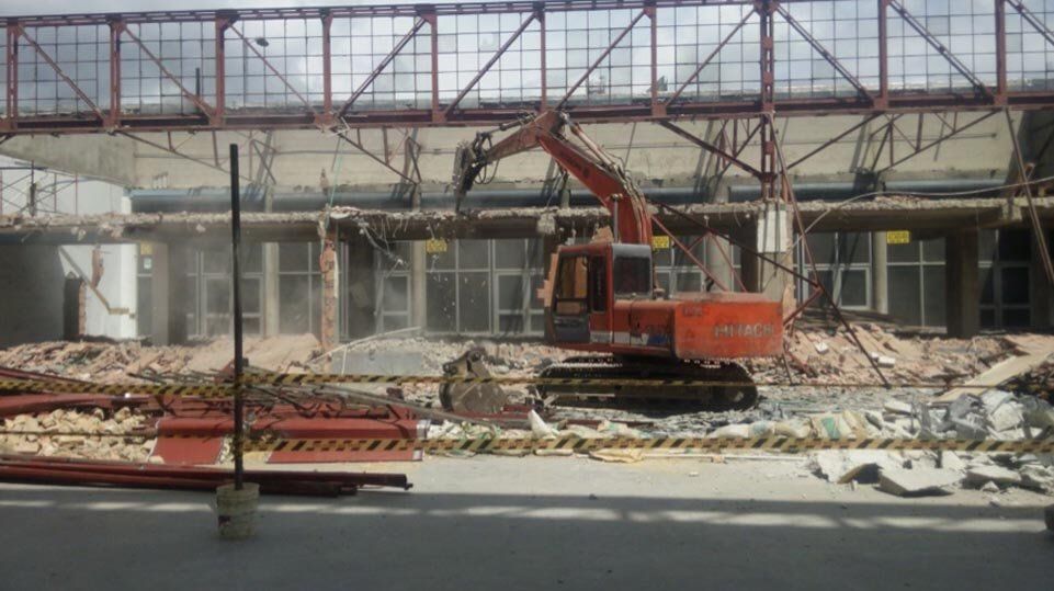 Una excavadora naranja demuele escombros junto a un edificio dañado en reconstrucción.