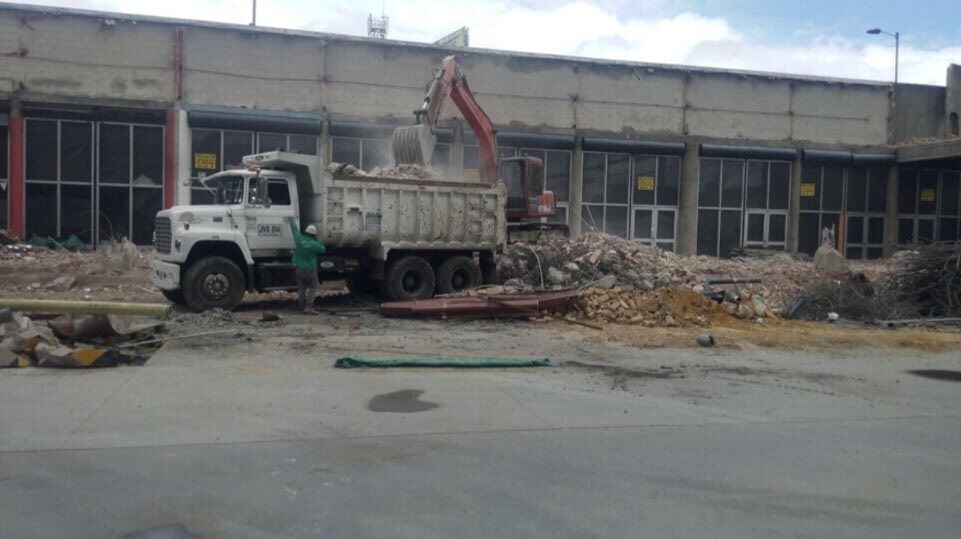 Un camión volquete y una excavadora entre los escombros de una demolición en las afueras de un edificio industrial dañado.
