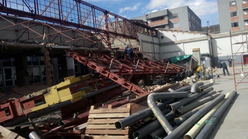 Estructura de acero rojo derrumbada en una obra en construcción, con tuberías y escombros esparcidos a plena luz del día.