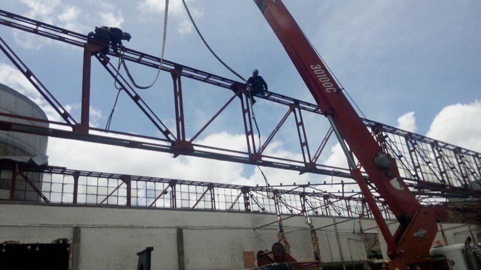 Trabajadores en una grúa levantando una gran estructura de acero sobre una obra en construcción bajo un cielo azul.