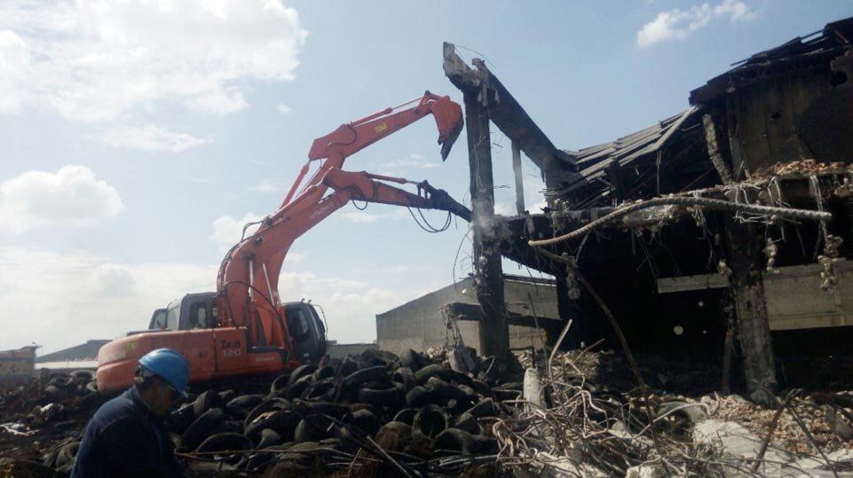 Una excavadora naranja demuele un edificio incendiado entre escombros, con un trabajador con casco azul en primer plano.