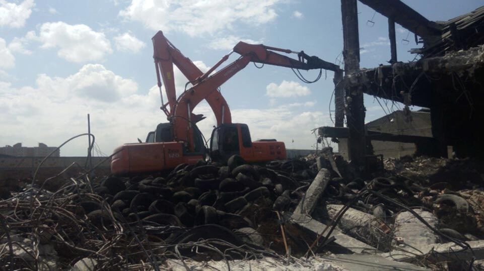Una excavadora naranja retira escombros en un sitio de demolición junto a un edificio dañado.