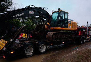 Una excavadora John Deere amarilla cargada en un remolque de plataforma junto a un camión rojo en una carretera mojada.