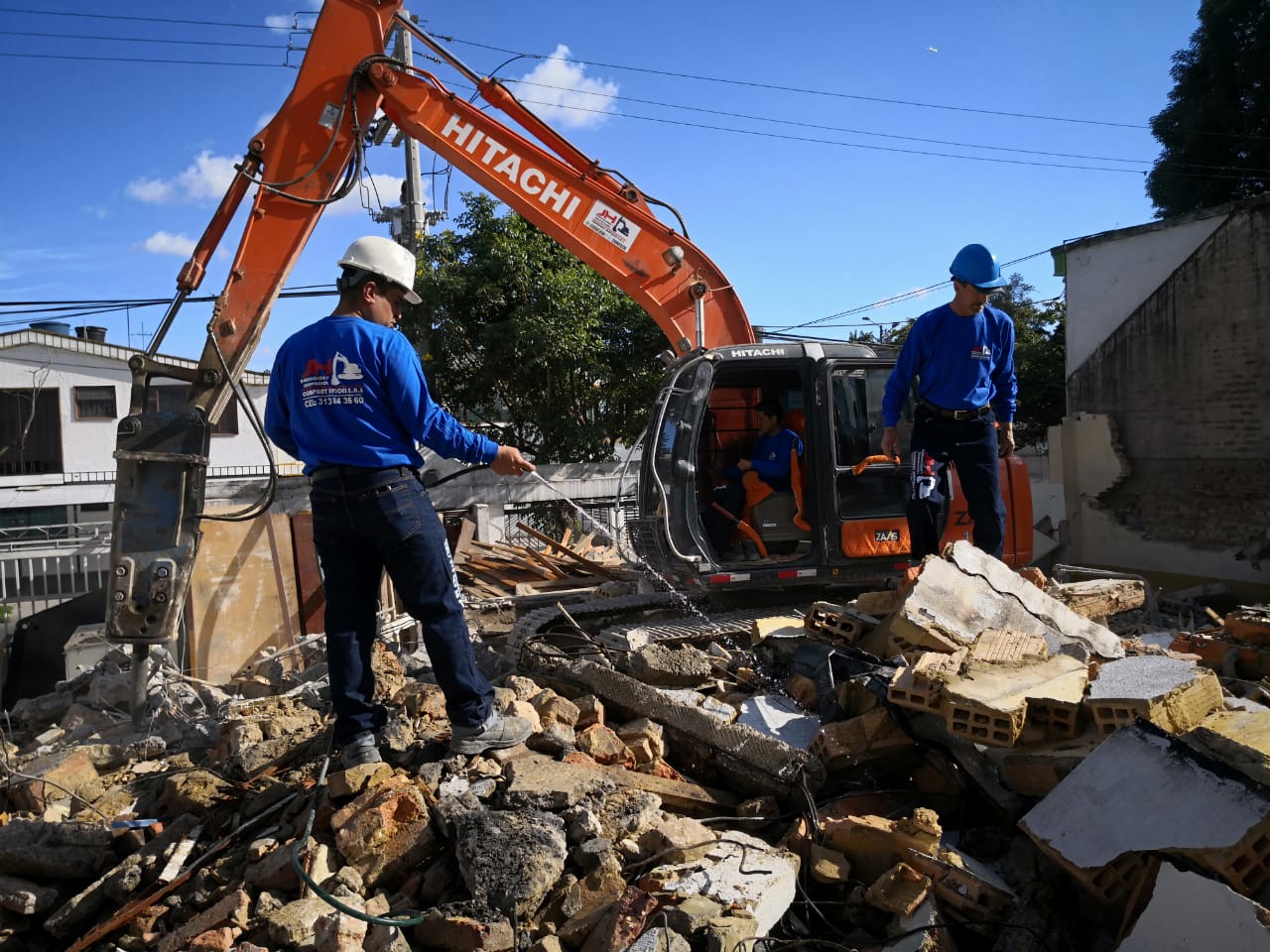 Excavadora retirando escombros en una obra de demolición con dos trabajadores con equipo de seguridad azul.