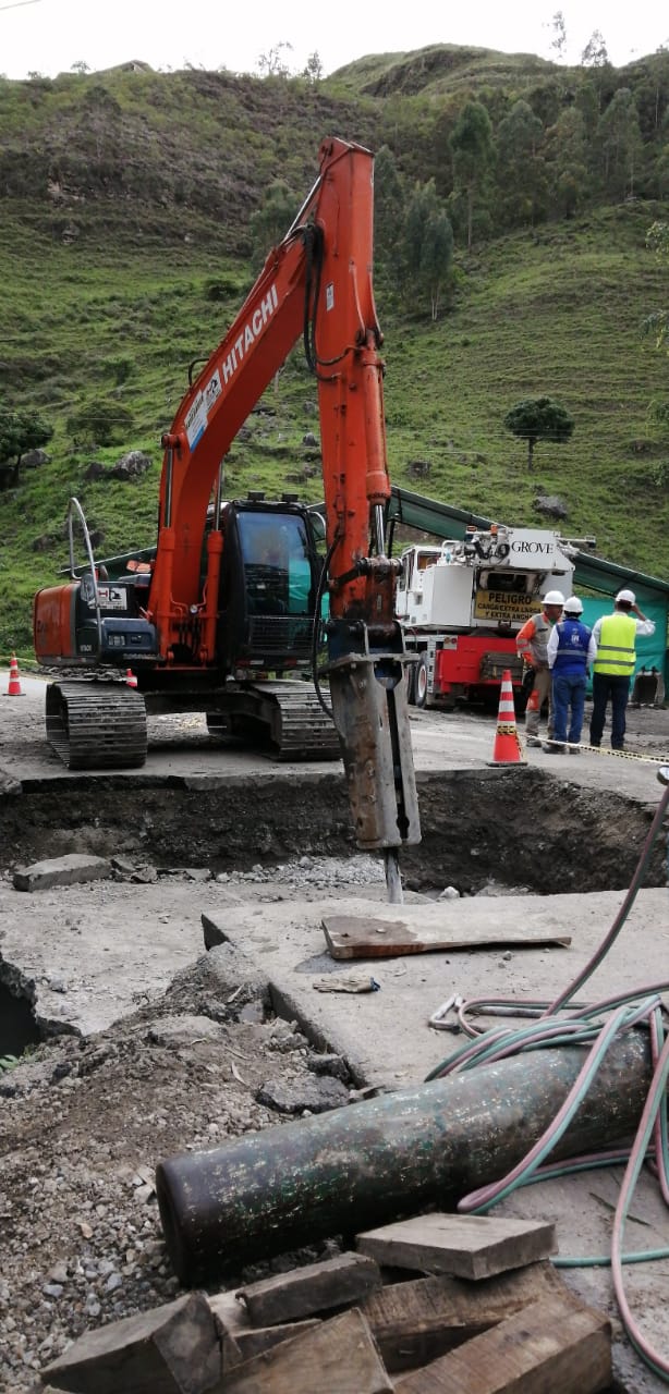 Excavadora naranja junto a trabajadores en una obra con pavimento roto y restos de maquinaria.