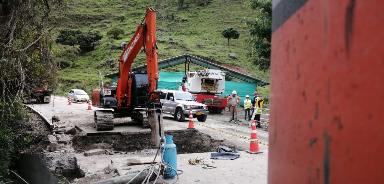 Excavadora naranja trabajando en un arcén embarrado cerca de coches y conos de tráfico en una zona verde de ladera.