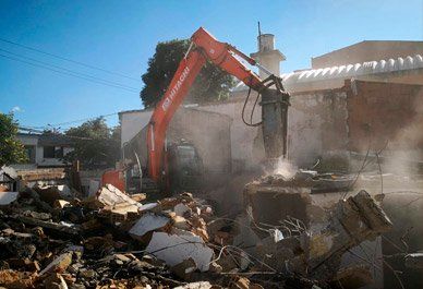 Una excavadora naranja demuele un edificio dañado entre escombros y polvo bajo un cielo azul despejado.