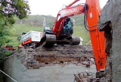 Excavadora y camión volquete trabajando en una carretera derrumbada junto a un terraplén erosionado.