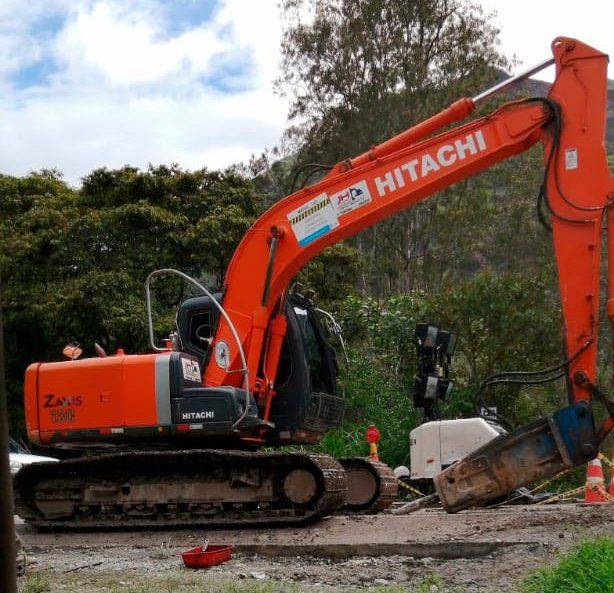 Excavadora Hitachi naranja en un terreno baldío levantando una pala cerca de árboles y cielo nublado.