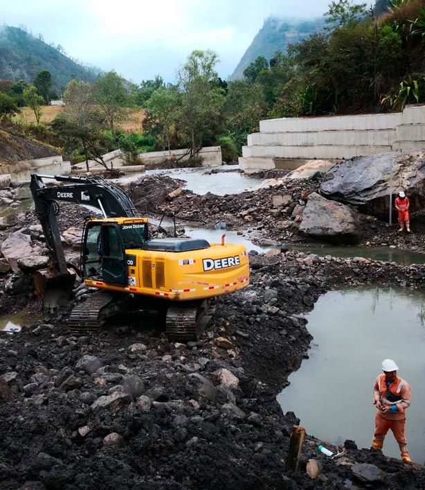 Una excavadora amarilla retira lodo y escombros cerca de un camino rocoso inundado, con trabajadores con cascos cerca.
