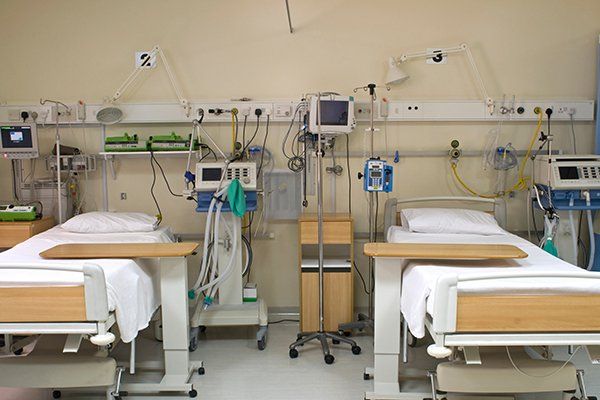 Two hospital beds in a medical room, with monitors and equipment mounted on the wall. Both beds are made and ready for patients.