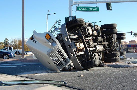 A white semi-truck overturned on a city street after a traffic accident. The sky is blue, and other vehicles are present.