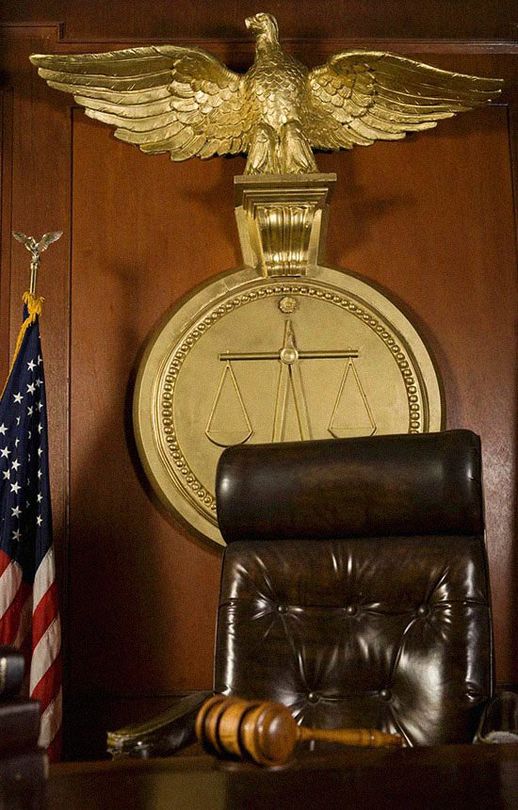 Judge's bench in a courtroom with a golden eagle, scales of justice, American flag, and gavel. A leather chair sits in front of the desk.