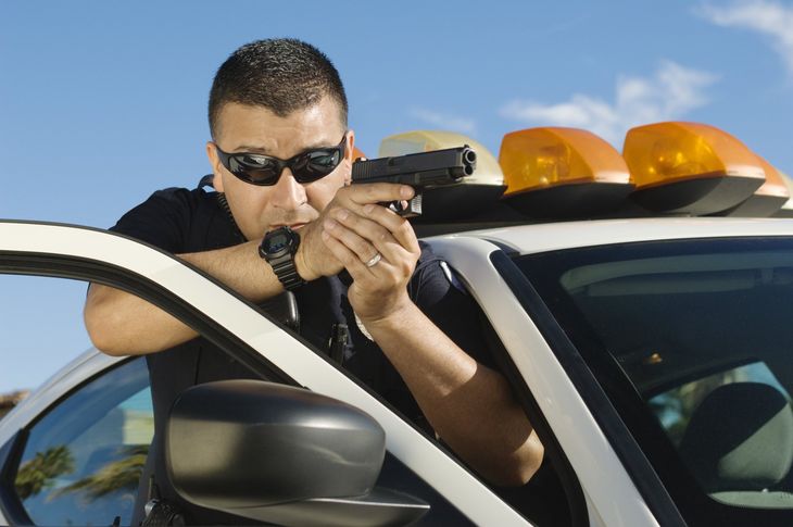 A police officer in sunglasses aims a handgun, leaning out of a patrol car. The car has flashing amber lights.