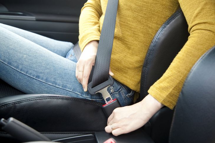 A person wearing a yellow sweater and jeans fastens a black seatbelt in a car.