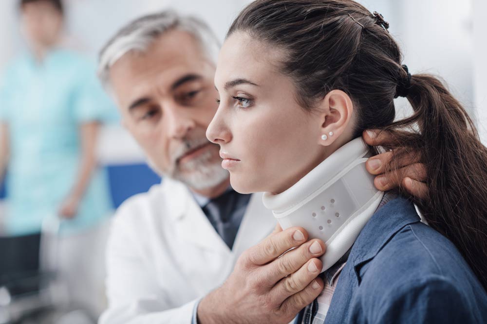 Doctor fitting a neck brace on a woman in a clinic setting. The woman has a pained expression while the doctor examines her neck.