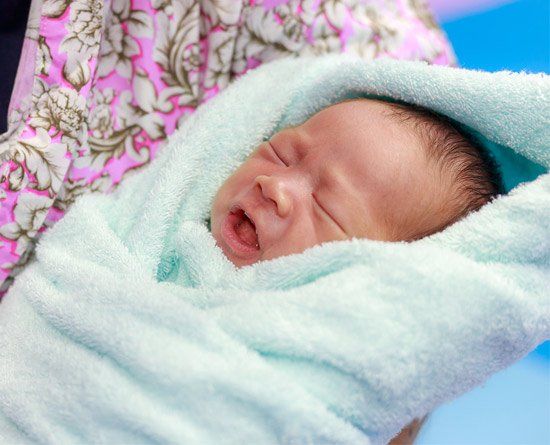 Newborn baby wrapped in a light blue towel, held by a person wearing a floral shirt. Baby is sleeping with mouth slightly open.