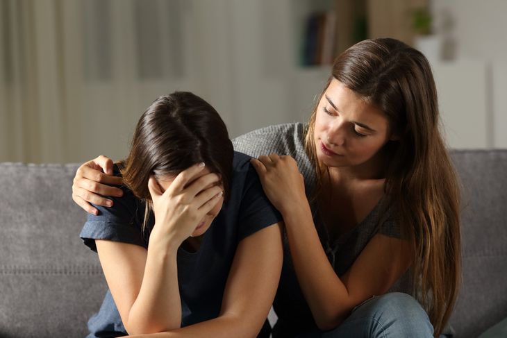 Woman comforting another woman who is crying, indoors on a couch. One has hand over face; other has arm around shoulder.