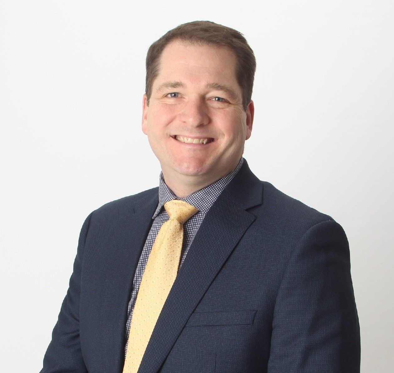 Man in navy suit, blue patterned shirt, and yellow tie smiles at the camera. Studio shot on a white background.