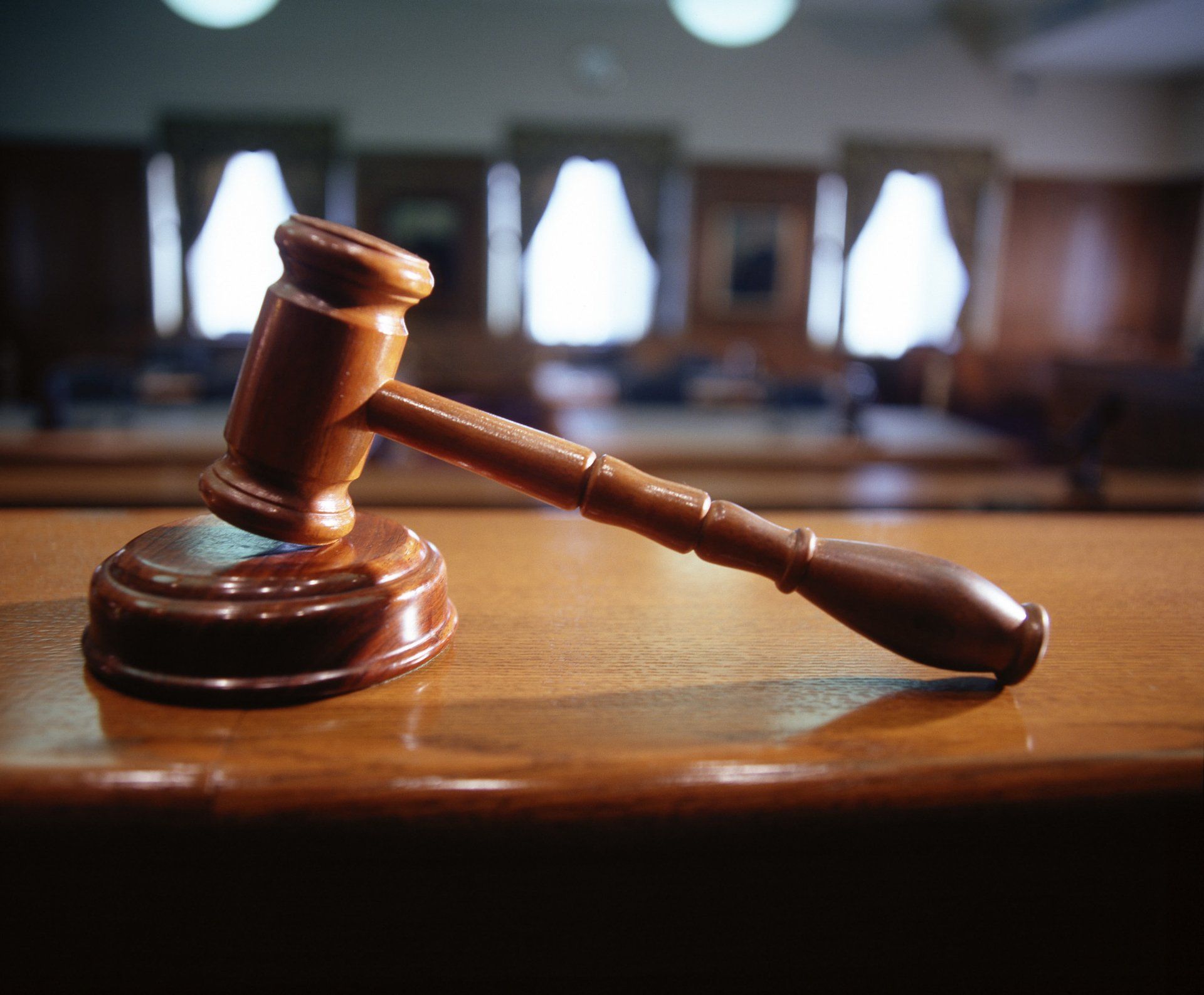 Wooden gavel resting on a table in a courtroom, with blurred windows and seating in the background.