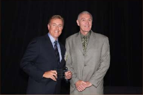 Two men in suits pose against a black backdrop. One holds an award, smiling; the other, older, stands beside him, smiling.