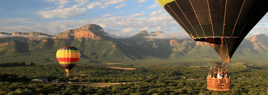 Two hot air balloons are flying over a lush green valley with mountains in the background.
