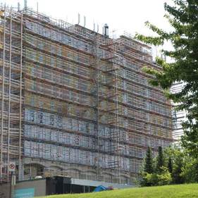Bâtiment en construction recouvert d'échafaudages, à côté d'arbres verts et d'un ciel nuageux
