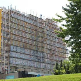 Bâtiment à plusieurs étages en rénovation, entouré d'échafaudages, à côté d'arbres et d'une zone herbeuse.