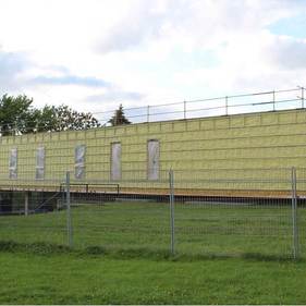 Bâtiment jaune derrière une clôture en grillage, avec de l'herbe devant et un ciel nuageux au-dessus.