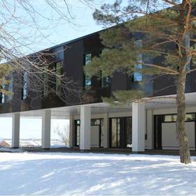 Bâtiment moderne et sombre sur pilotis, entouré de neige et d'arbres dénudés.