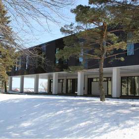 Bâtiment moderne et sombre aux grandes fenêtres, se détachant sur un paysage enneigé et encadré par des arbres.