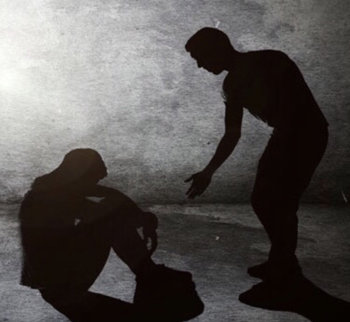 Silhouette of a person comforting another, offering a hand. Both are in a dimly lit space.
