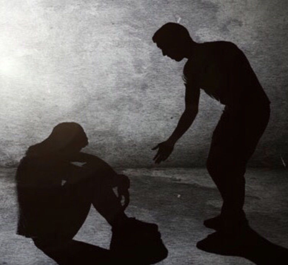 Shadow of one person offering a hand to another seated person, both against a textured wall.